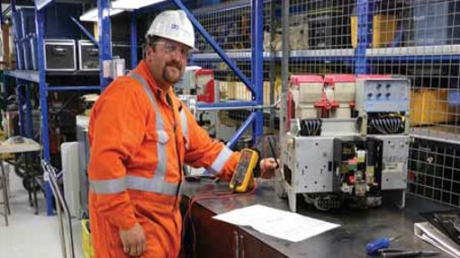 Dynamo electrician Ethan rebuilding a circuit breaker. The location is the maintenance shop in Saskatoon Sk. The employee is wearing a hard hat coveralls and protective eyewear.