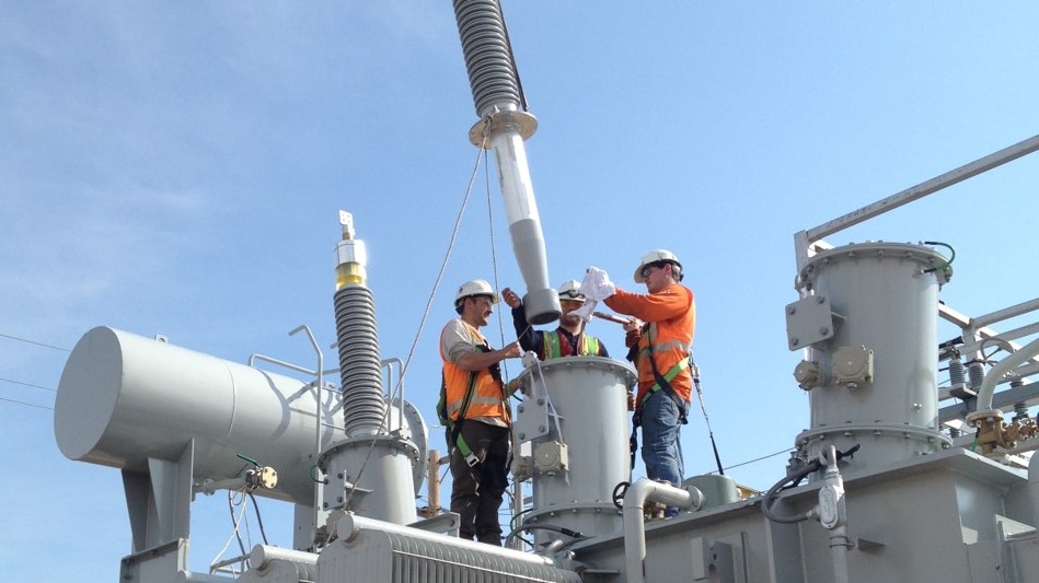 Dynamo construction electricians installing electrical insulators on top of a transformer bushing. Location is a substation. Employees are wearing a hardhats gloves and fall arrest harness.