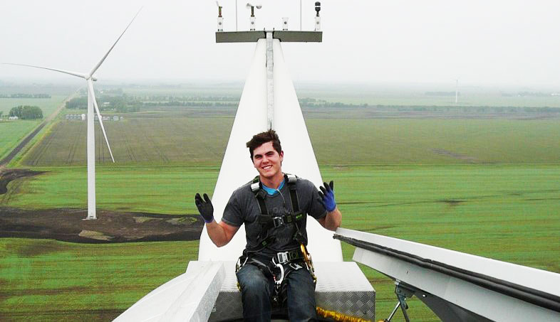 Dynamo employee sitting on the top of a wind turbine. The location is a field of wind turbines. The employee is wearing gloves and fall protection harness.