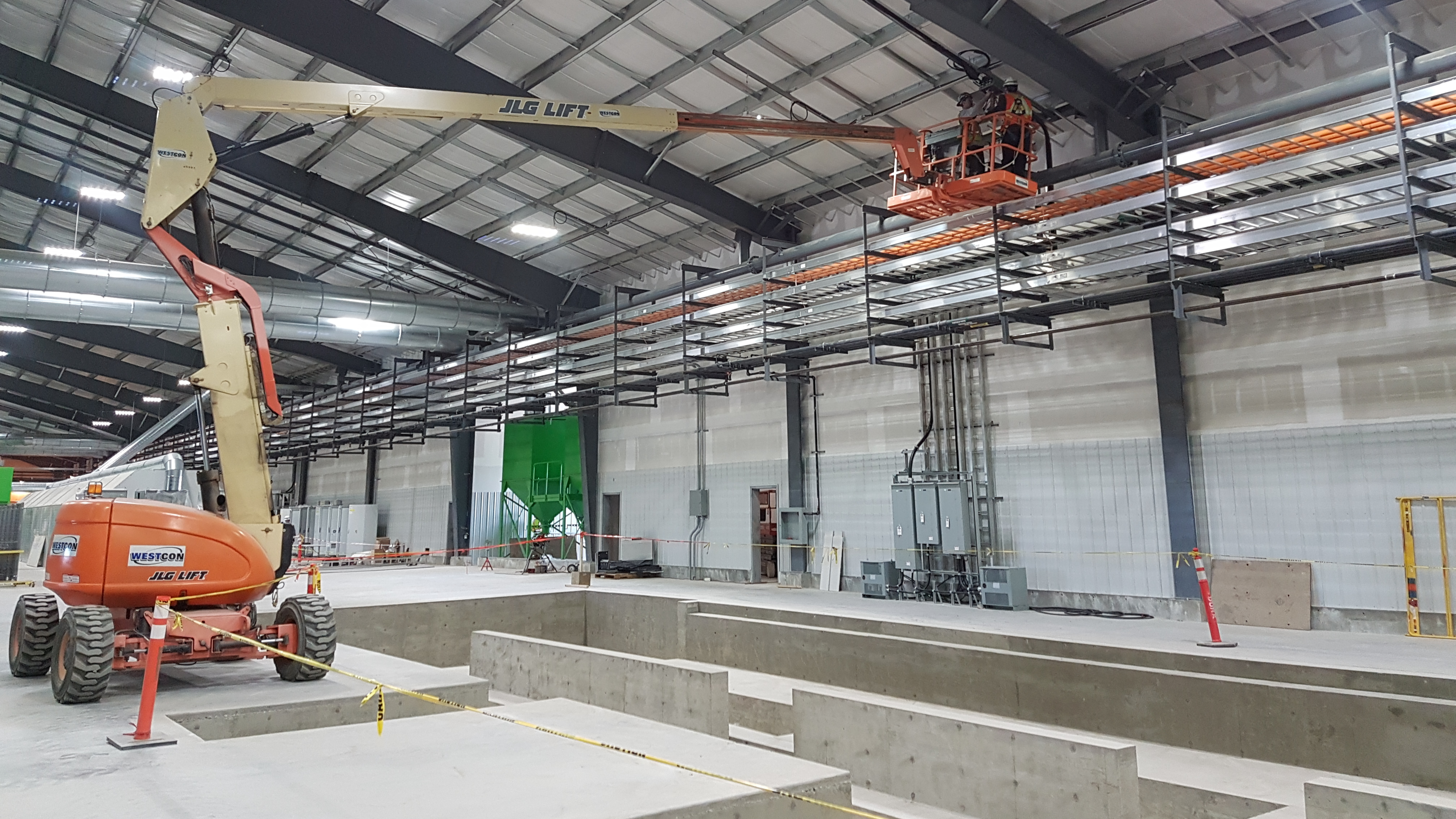  Dynamo employees on a boom lift high in the rafters runing electrical wire for an industrial building. All two employees are wearing Personal Protective safety equipment and fall arrest harasses.