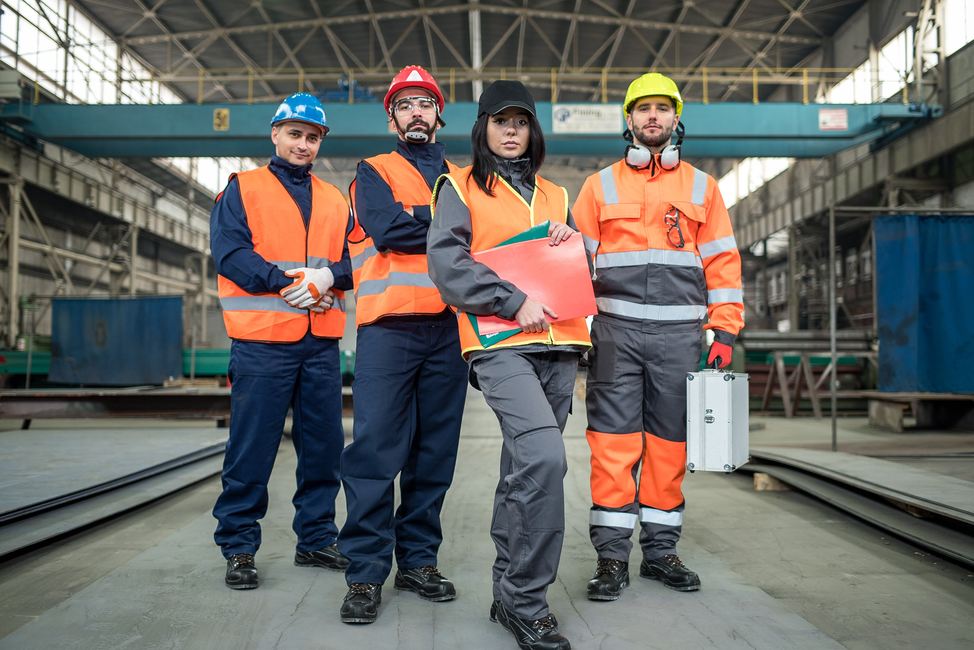   Four workers of different sex and race in a manufacturing warehouse. The employees are all wearing personal protective equipment.